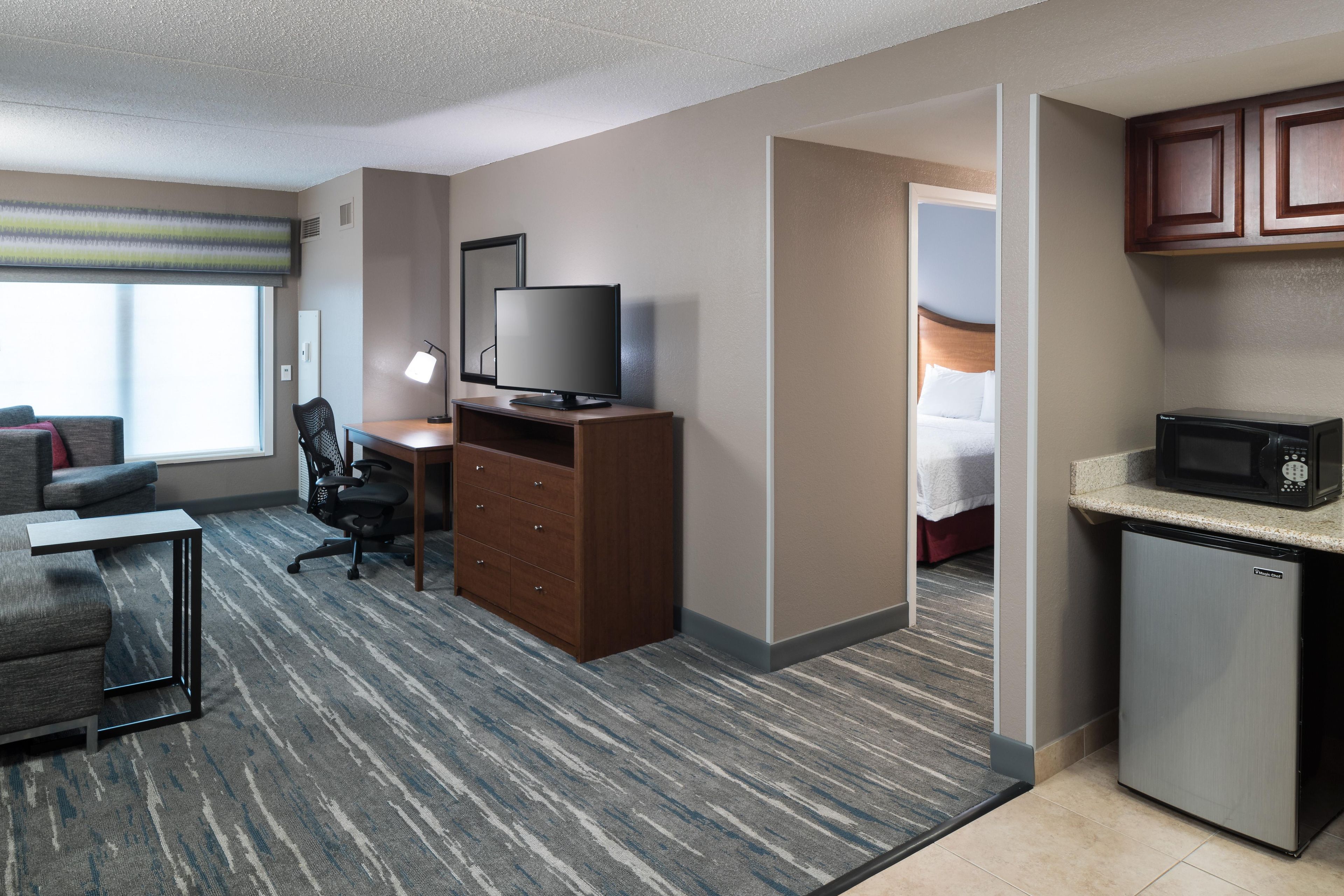 A hotel room with a desk, chair, television, minibar, and a view of the bed.