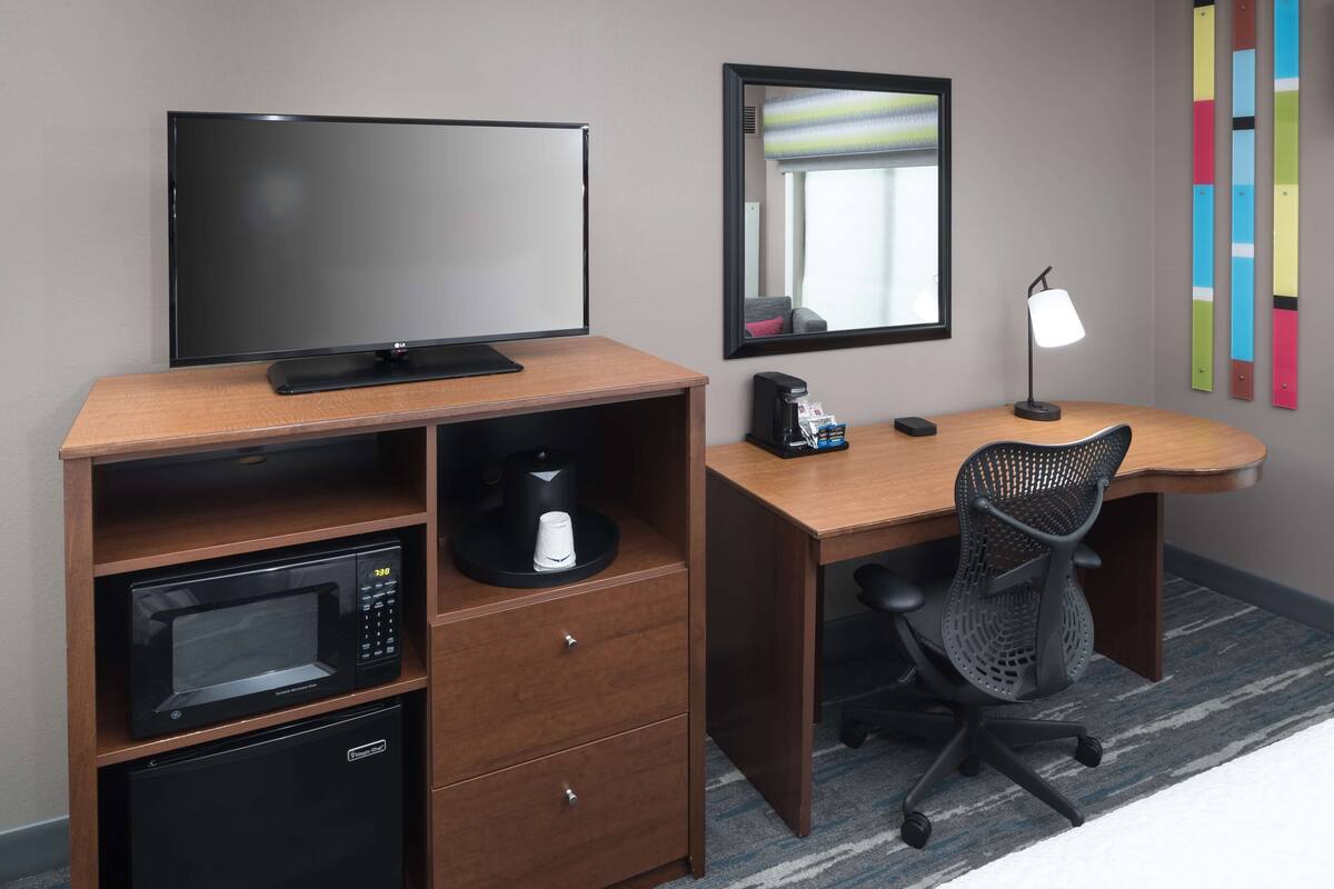 A hotel room with a wooden desk, a black office chair, a flat-screen TV, a microwave, a coffee maker, and a mirror.