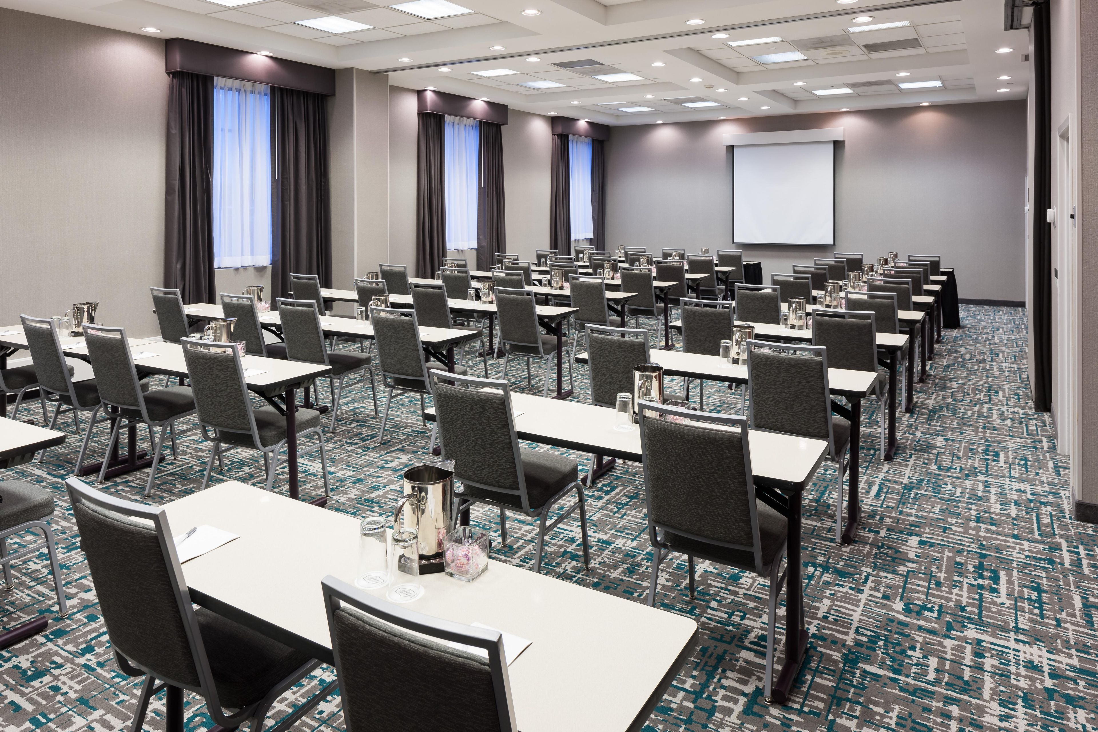 Ein Konferenzraum mit mehreren Tischen und Stühlen, einer Projektionsleinwand und einem gemusterten Teppich.