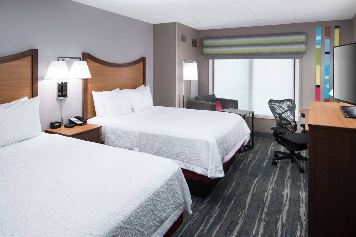 A hotel room with two beds, a desk, a chair, and a TV.