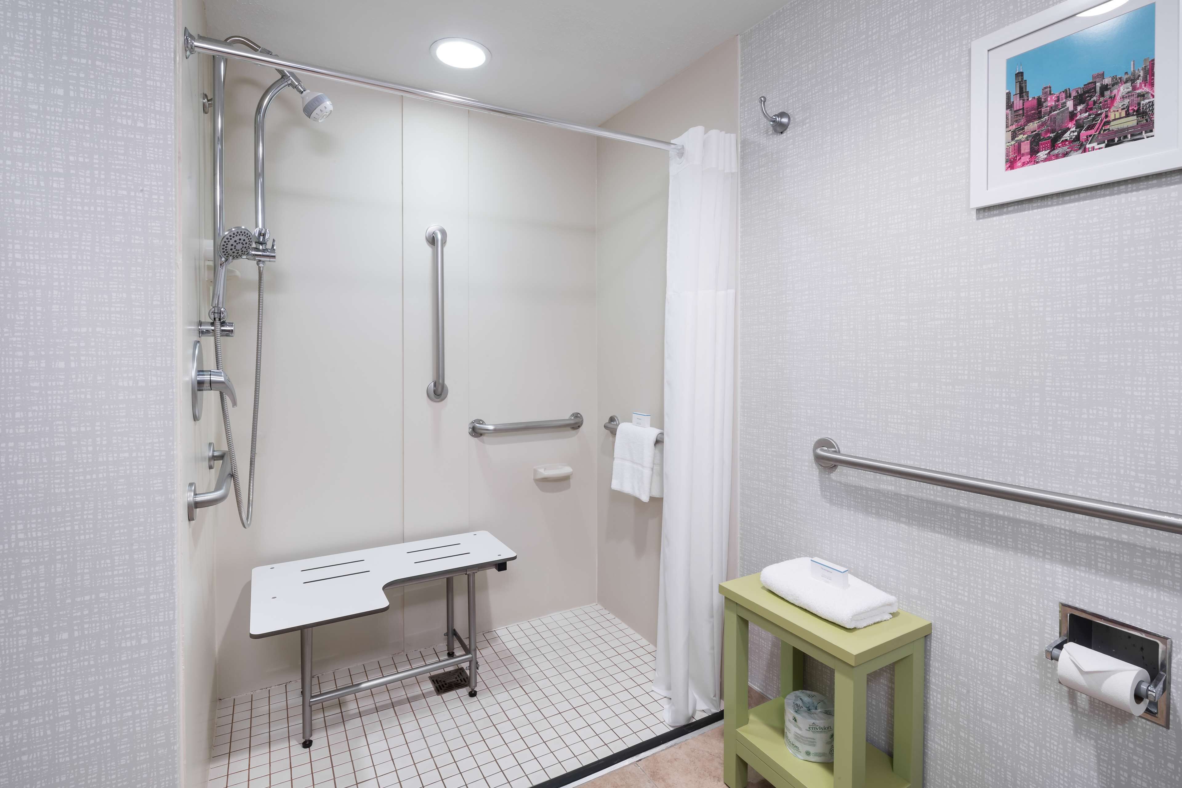 A modern bathroom with a shower, grab bars, a white sink, and a green stool.