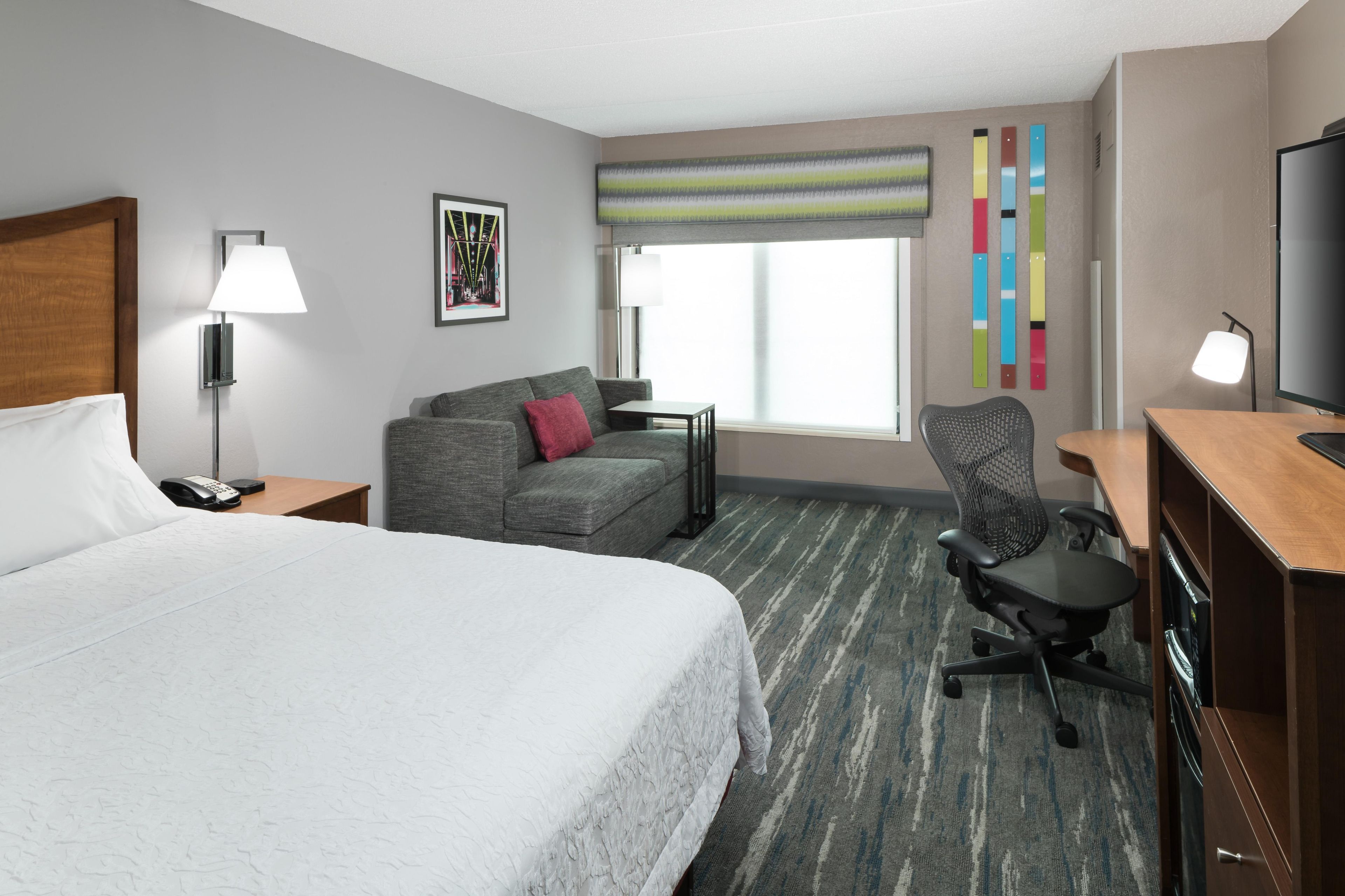 A hotel room with a large bed, a gray sofa, a desk with a chair, a television, and a window with a view.