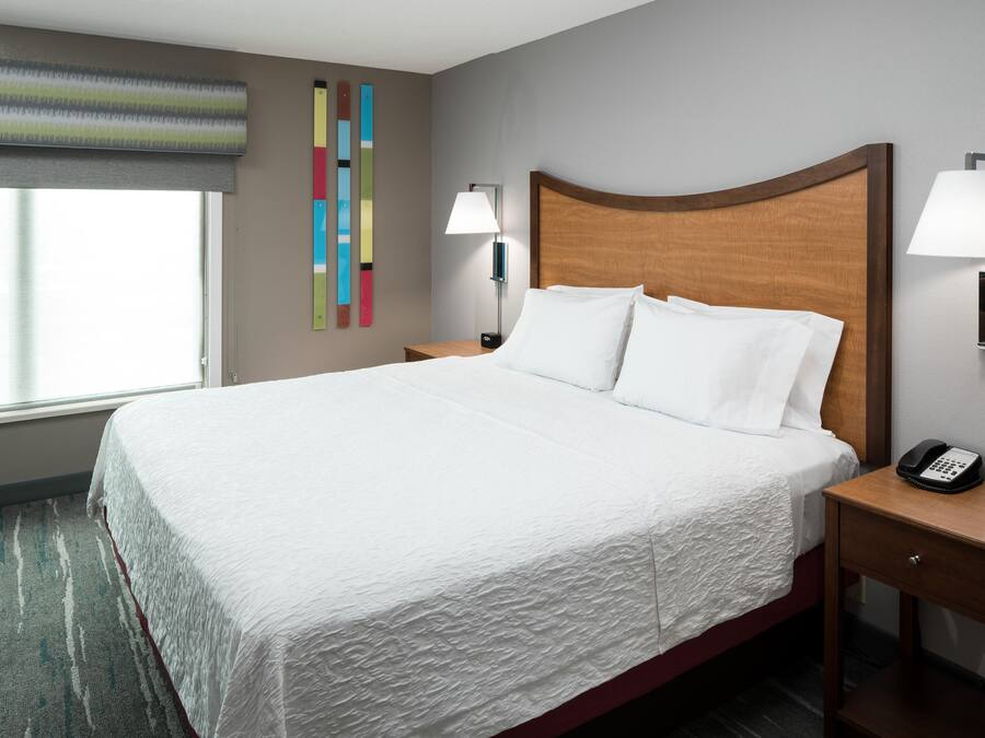 Una habitación de hotel con una cama grande, dos mesitas de noche con lámparas, un teléfono en la pared y una ventana con persianas.