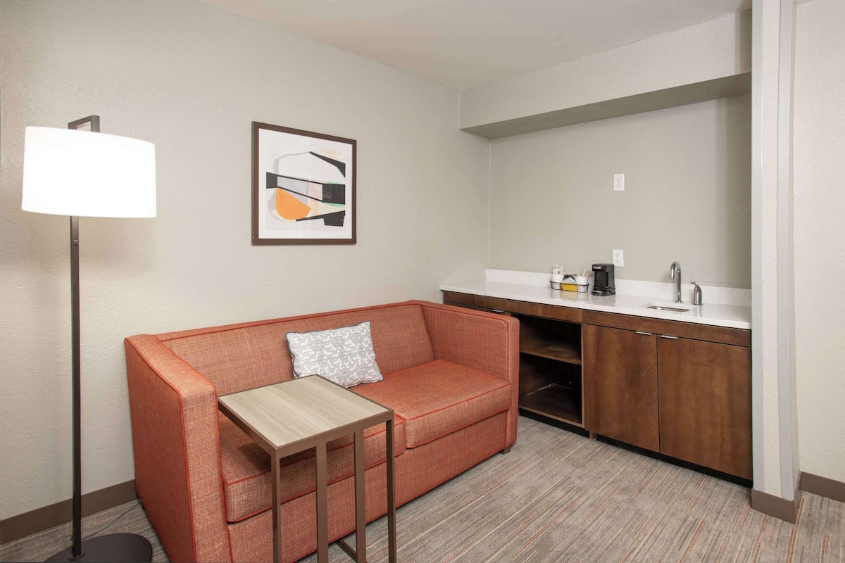 A compact hotel room with a sofa, a small table, a lamp, a framed abstract artwork, and a kitchenette.