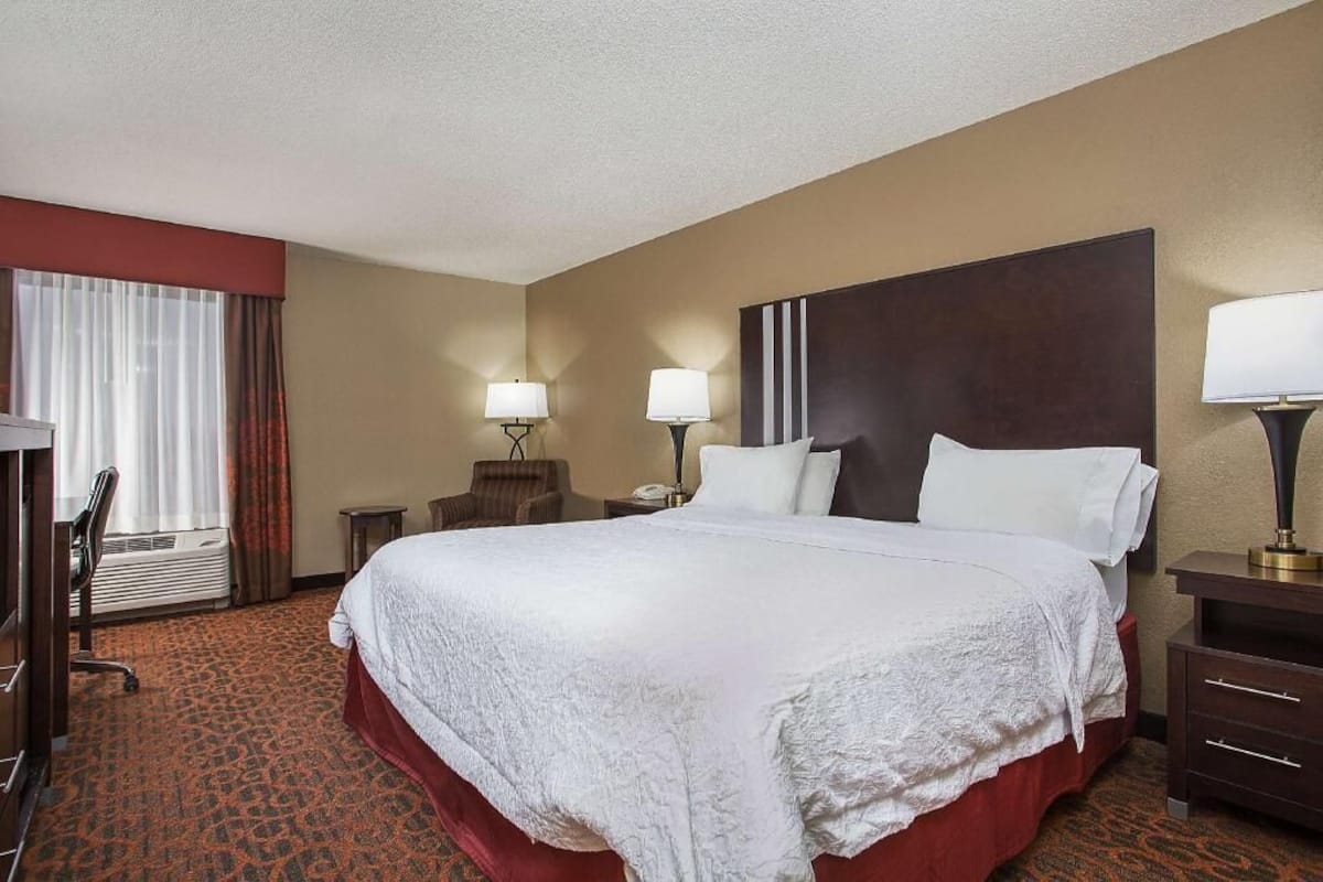 A hotel room with a large bed, two bedside tables with lamps, a chair, a desk, and a window with curtains.