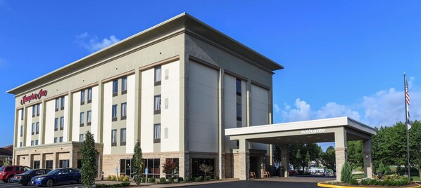 A modern hotel with a flat facade, a covered entrance, and a parking lot with several cars.