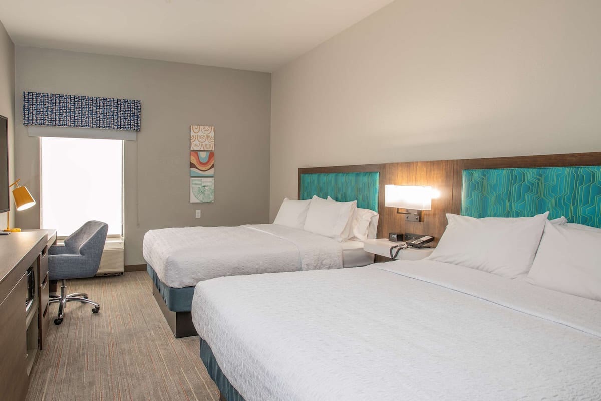 A hotel room with two beds, a desk, a chair, and a window.