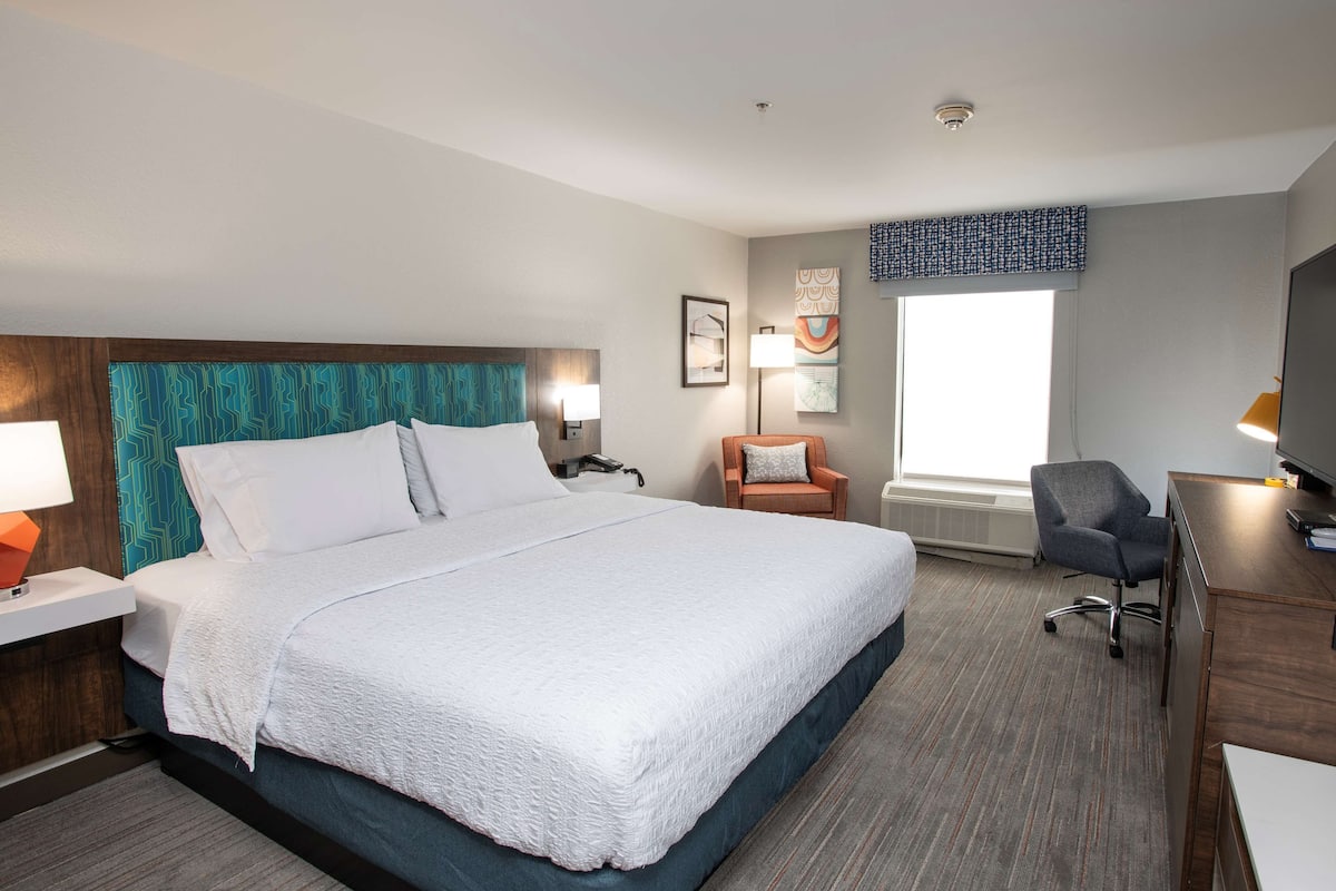 A hotel room with a large bed, a desk, a chair, a lamp, and a window.