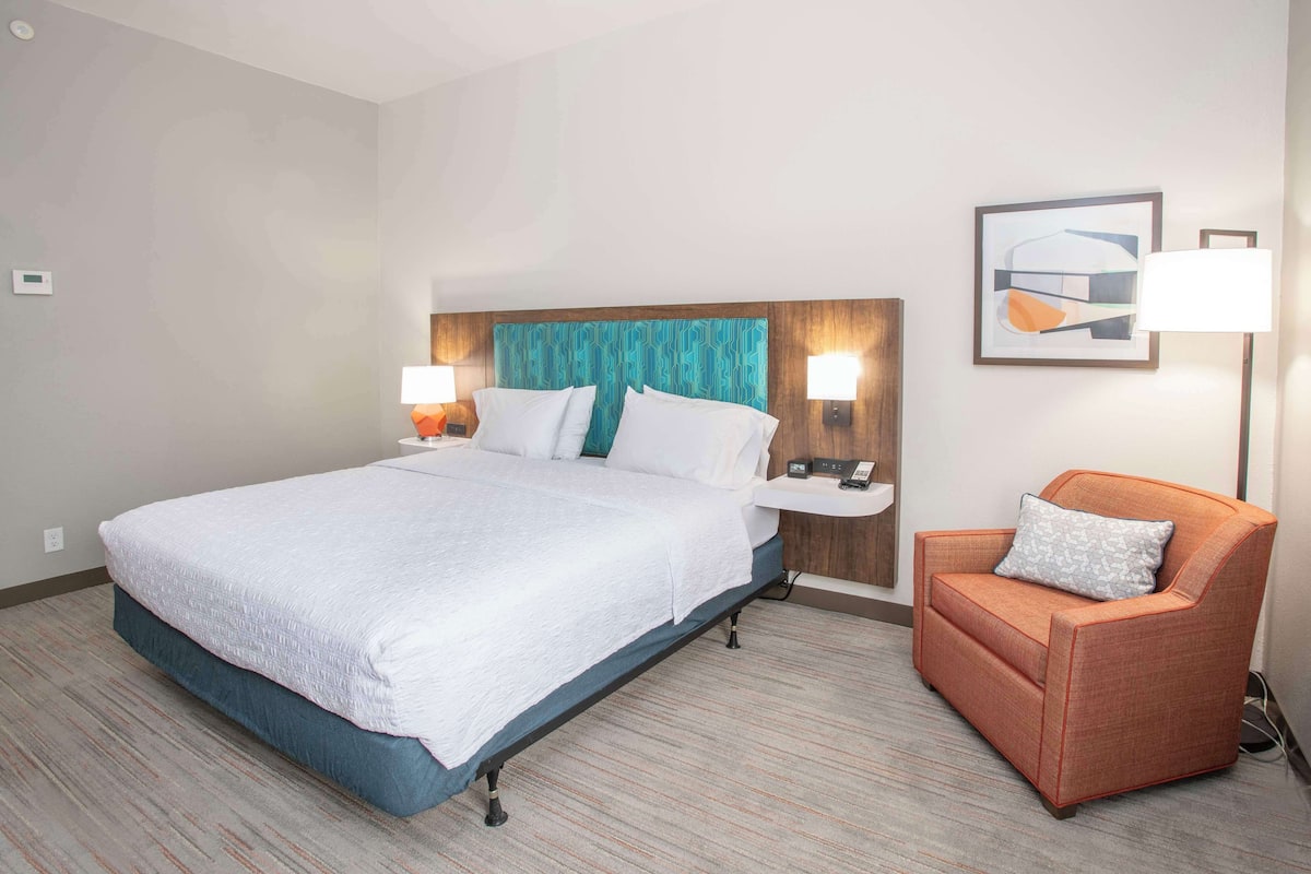 A hotel room with a large bed, a nightstand, a chair, and a wall-mounted lamp.