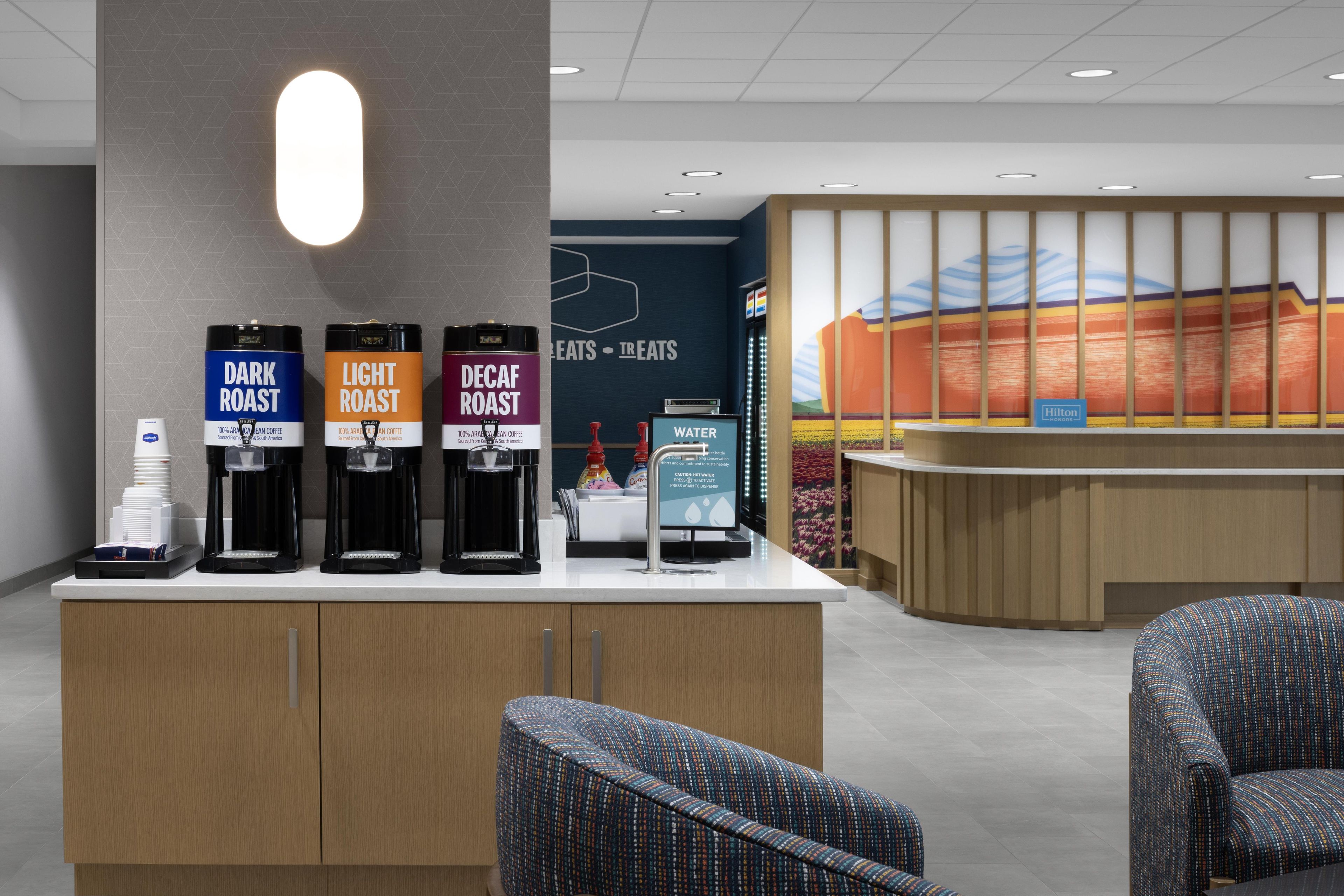 A hotel reception area with a coffee station featuring three coffee options: Dark Roast, Light Roast, and Decaf Roast. There are two chairs and a water dispenser.