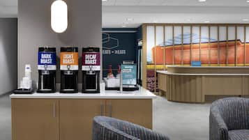 A hotel reception area with a coffee station featuring three coffee options: Dark Roast, Light Roast, and Decaf Roast. There are two chairs and a water dispenser.
