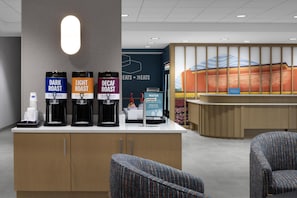 Un espace d’accueil d’hôtel avec un coin café proposant trois types de café : café torréfié foncé, café torréfié léger et café décaféiné. On y trouve deux chaises et un distributeur d’eau.