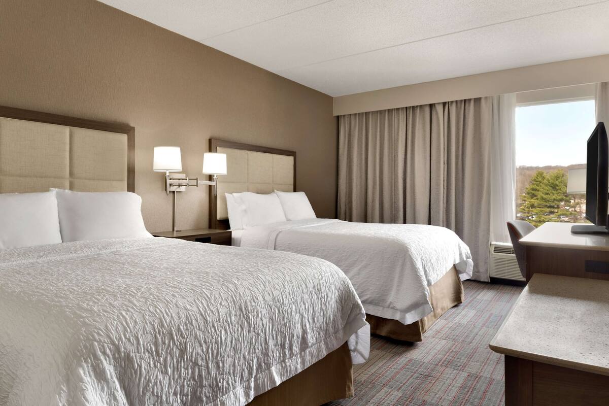 A hotel room with two beds, a desk, a television, and a window with curtains.