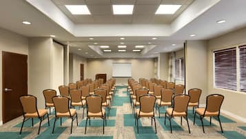 Un salón de conferencias con filas de sillas, una pantalla de proyector y piso alfombrado.