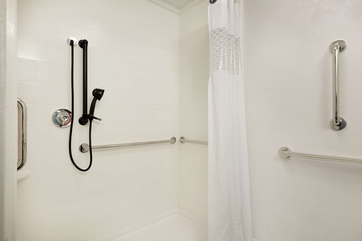 A shower area with white tiled walls, a handheld showerhead, a glass shower door, and a grab bar on the right side.