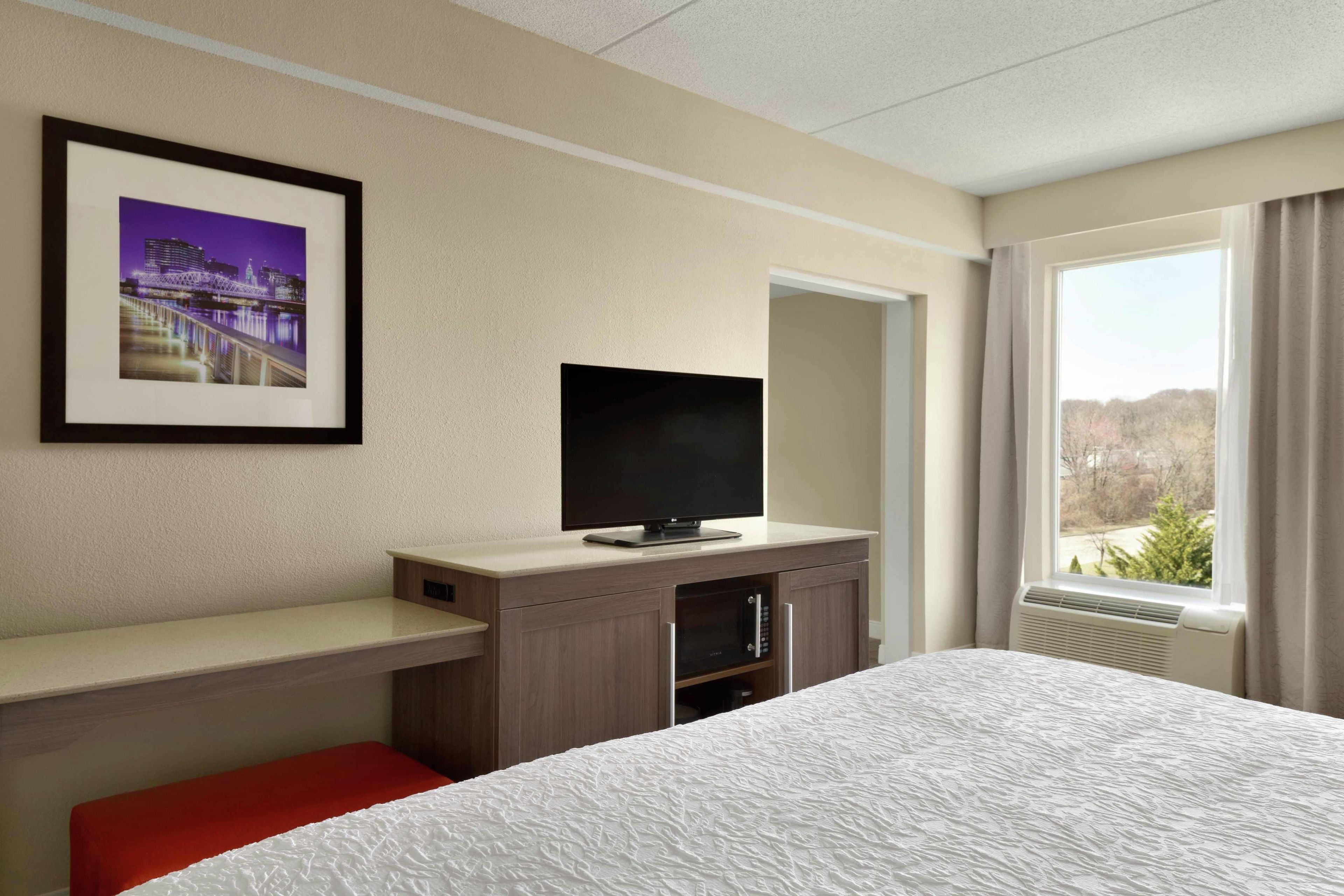 Habitación de hotel con cama, televisión sobre un soporte de madera, un cuadro en la pared y ventana con vistas.