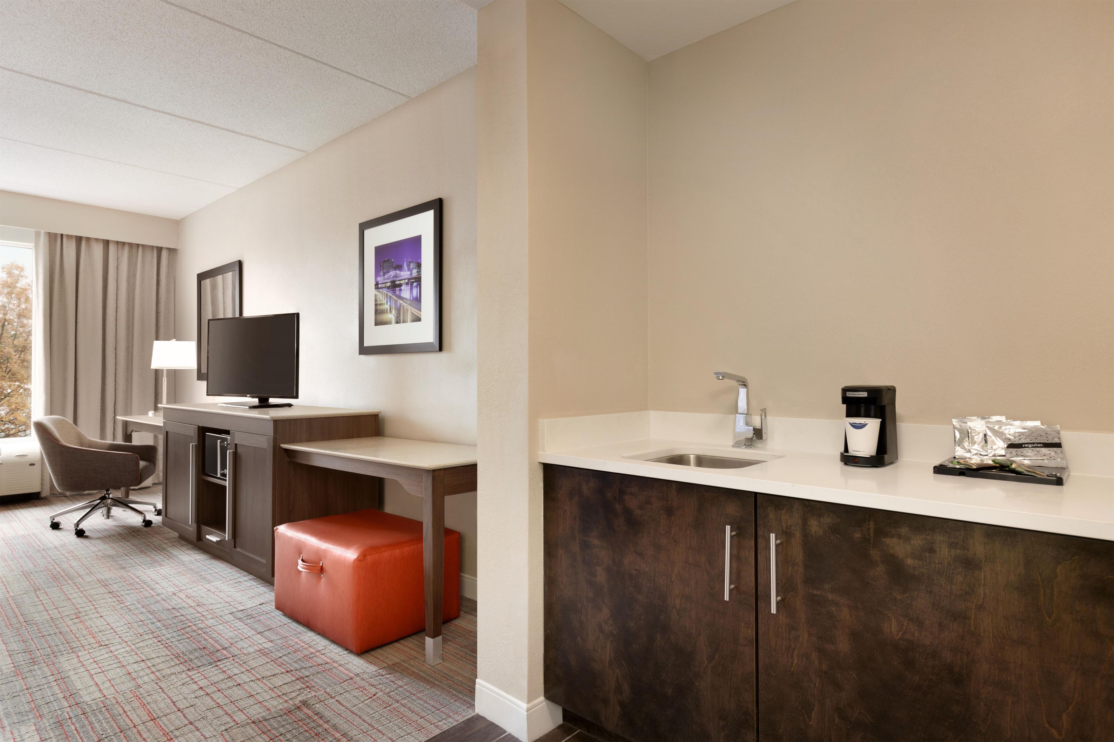 A hotel room with a desk, chair, television, and a kitchenette with a coffee maker and tea bags.