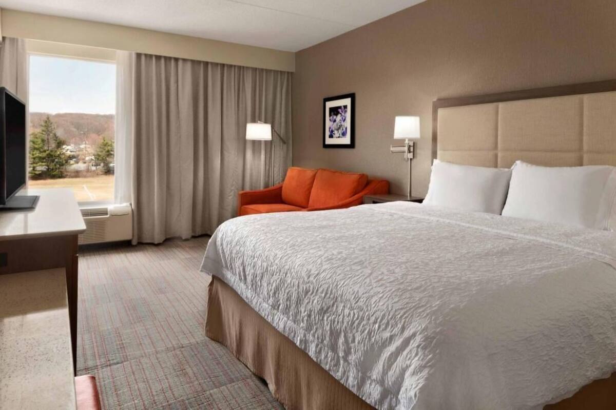 A hotel room with a large bed, a desk, a television, a window with curtains, and a framed picture on the wall.