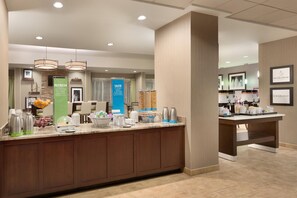A hotel breakfast area with a counter, coffee makers, fruit bowls, and a blue sign. - Hampton Inn Denville/Rockaway/Parsippany (Denville)