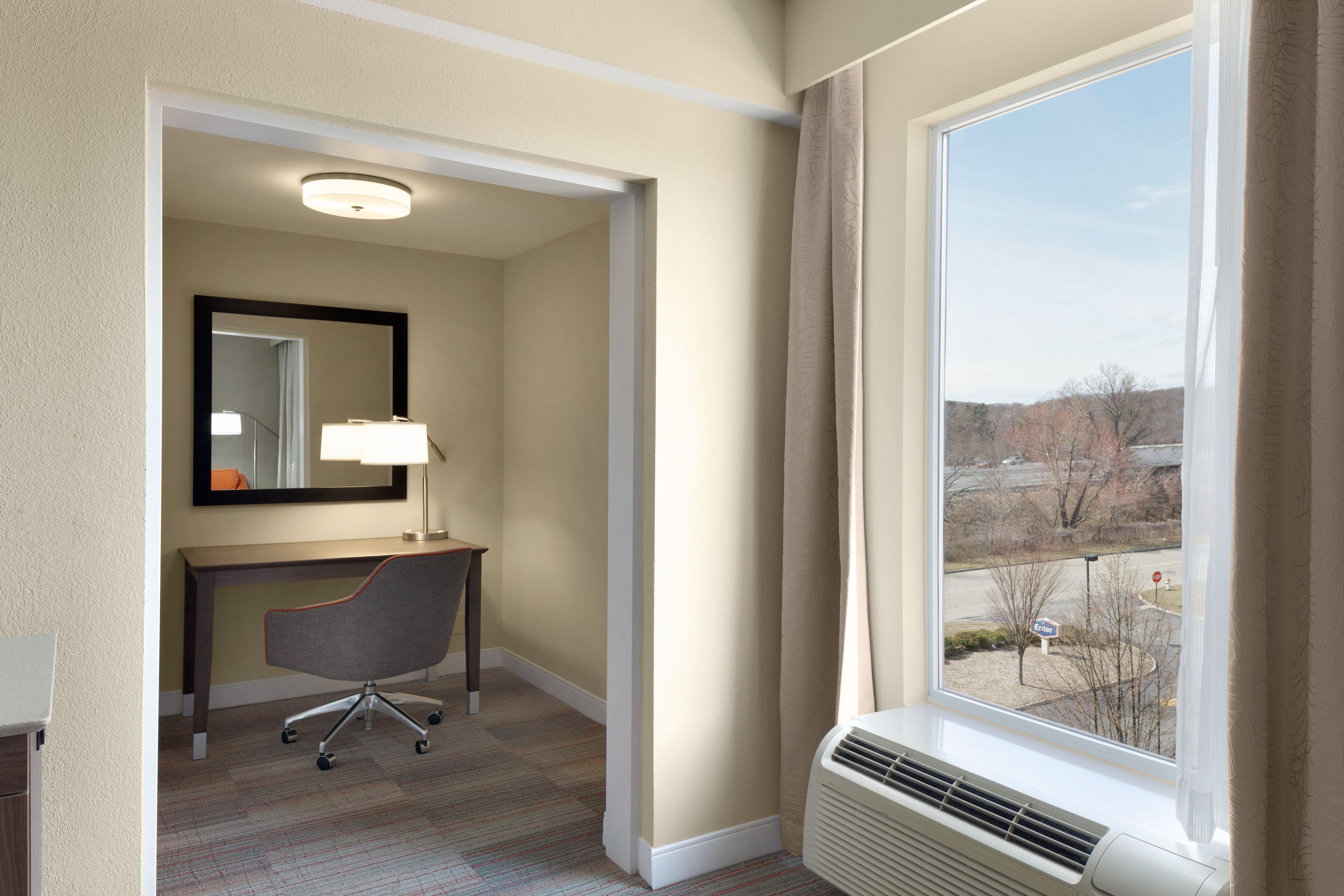A hotel room with a desk, chair, lamp, and a large window with a view of the outside.