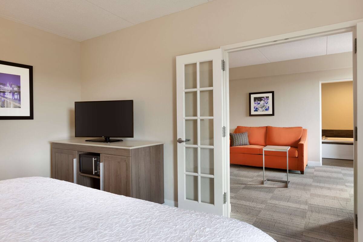 A hotel room with a bed, a TV on a wooden cabinet, a glass door leading to a living area with a sofa and a picture on the wall.
