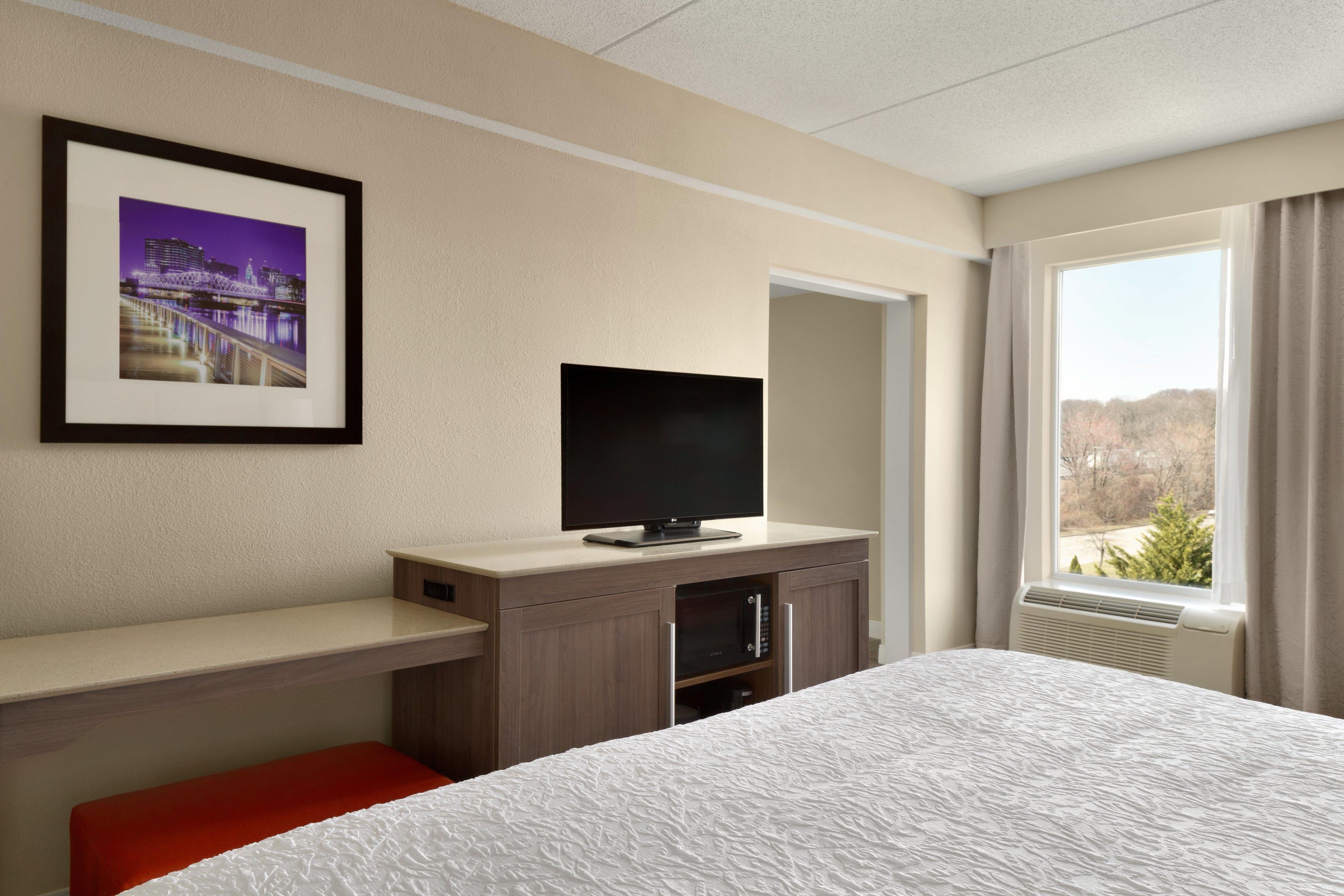 Une chambre d’hôtel avec un lit, un téléviseur posé sur un meuble en bois, un tableau encadré au mur et une fenêtre donnant sur un paysage.