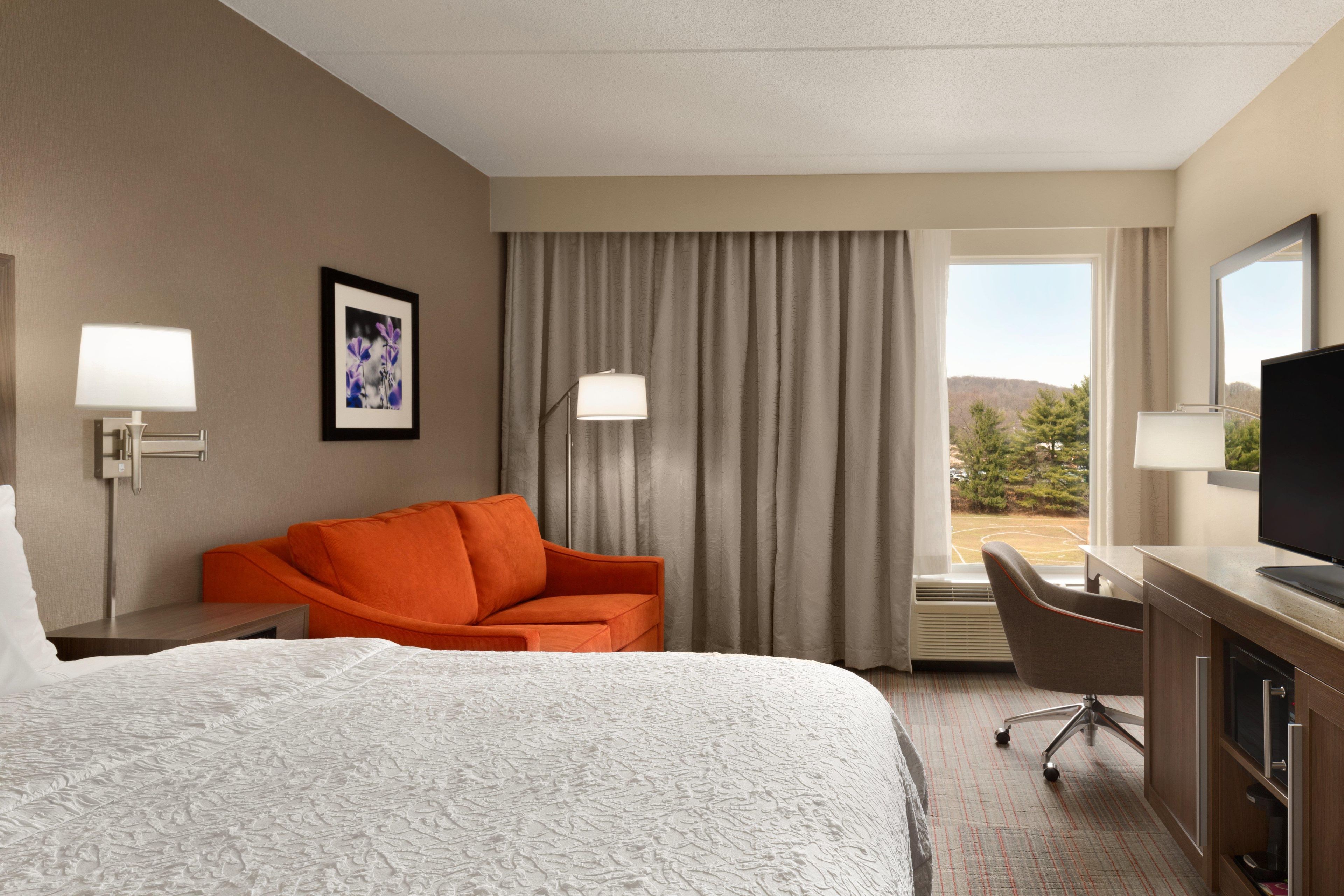 A hotel room with a large bed, an orange armchair, a desk with a computer, and a window with a view of trees.