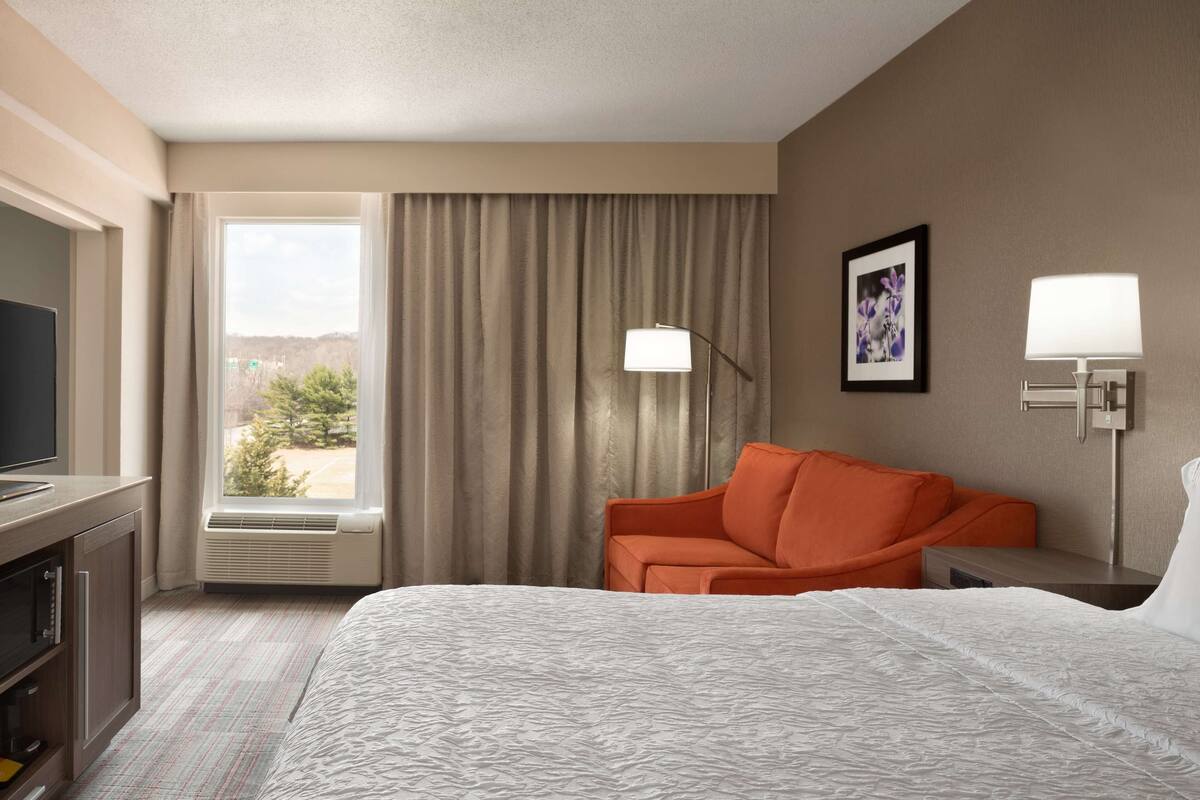 A hotel room with a bed, a television, an air conditioning unit, a sofa, and a window with curtains.