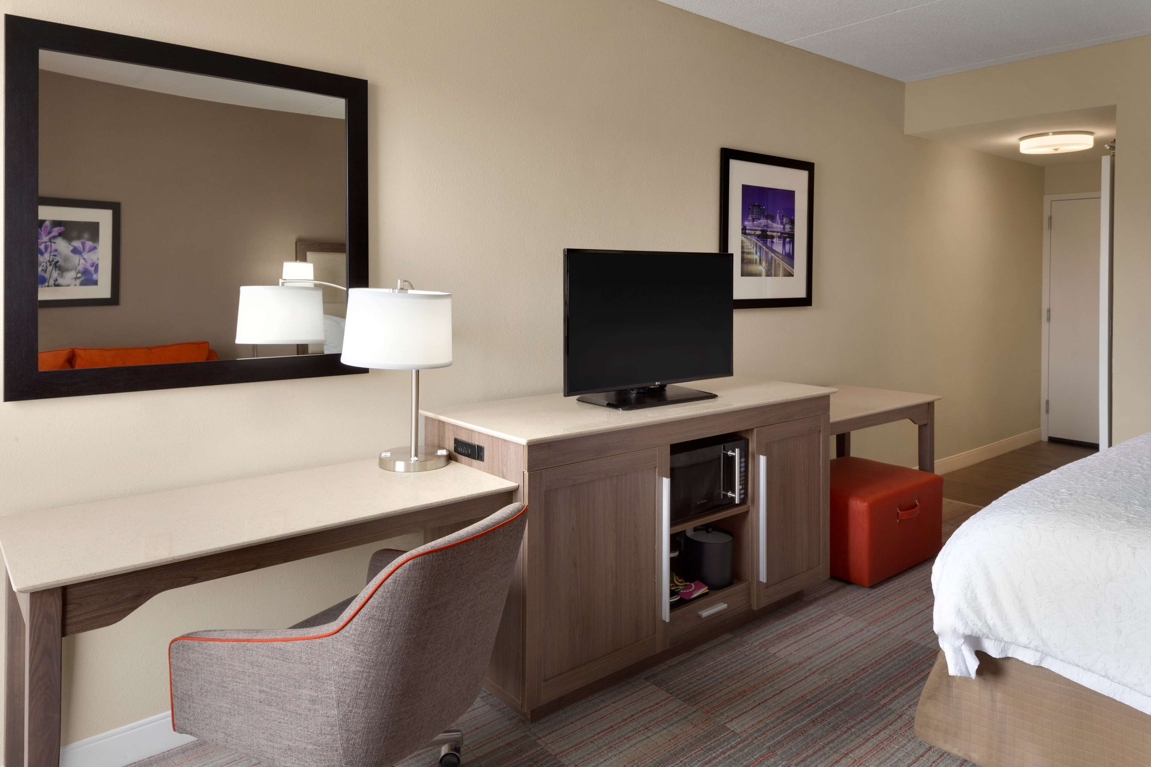 Habitación de hotel con cama, escritorio con televisor, silla, espejo y maleta roja.