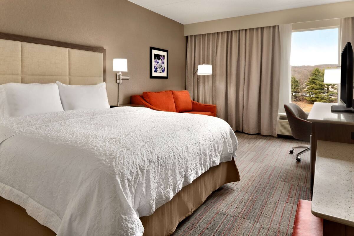 A hotel room with a large bed, a desk with a chair, a television, and a window with curtains.