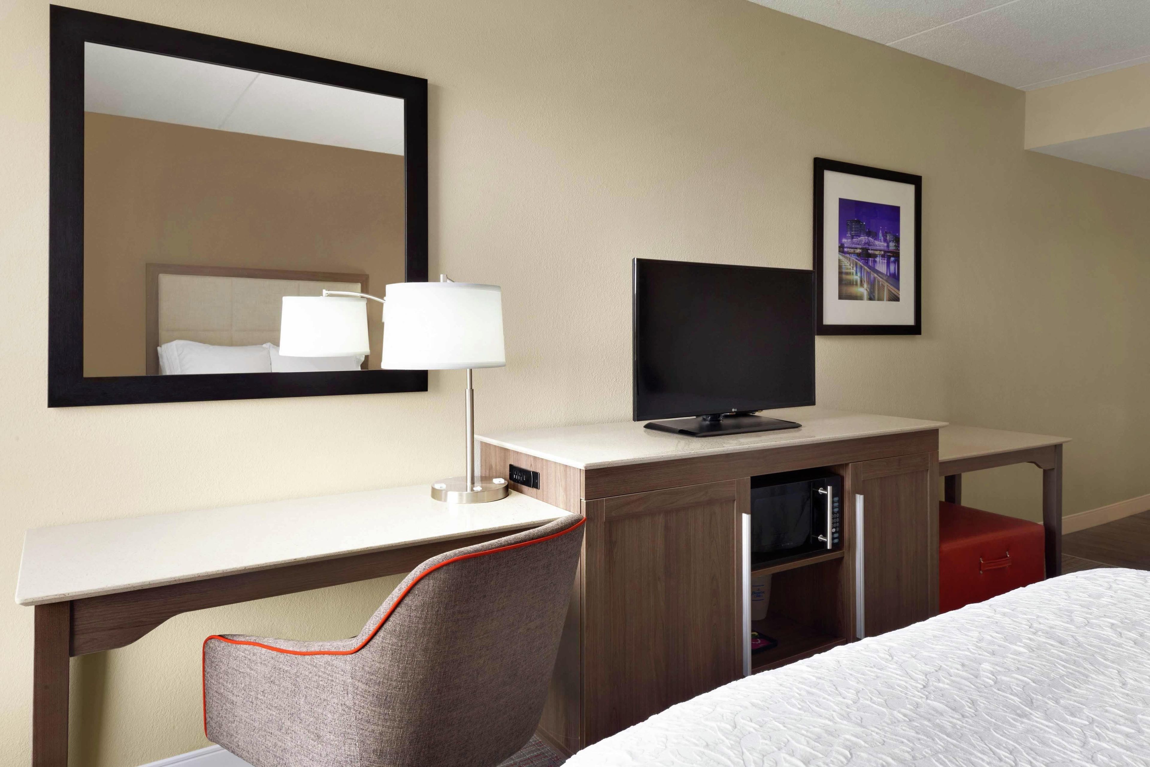 A hotel room with a bed, a desk with a television, a chair, a mirror, and a framed picture on the wall.