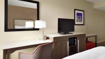 A hotel room with a bed, a desk with a television, a chair, a mirror, and a framed picture on the wall.
