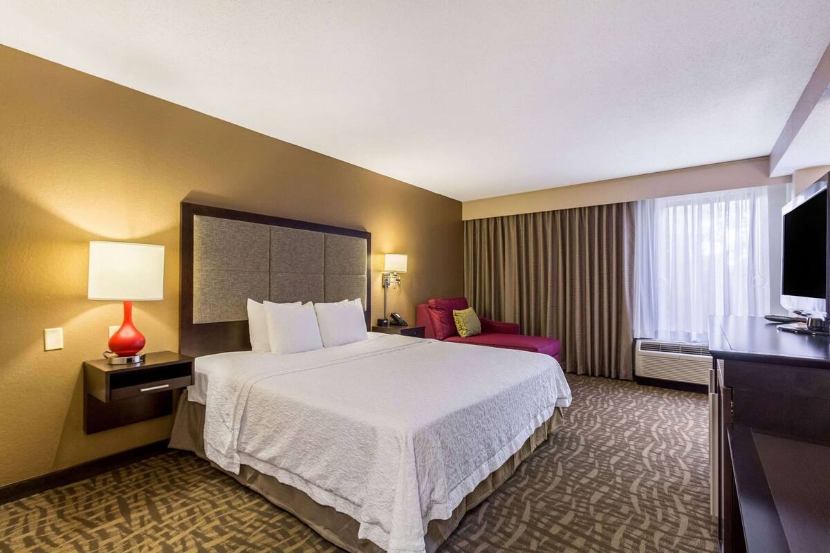 A hotel room with a large bed, a desk, a chair, a TV, and a window with curtains.