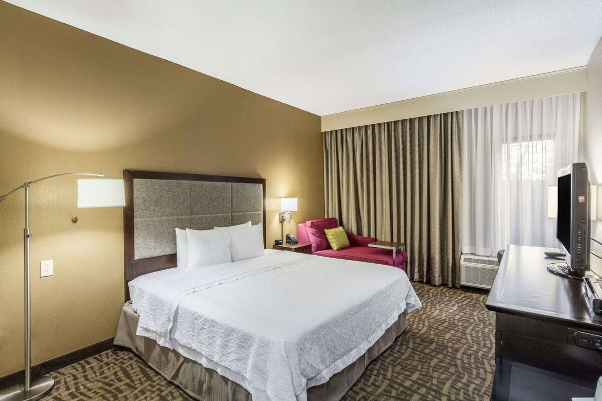 A hotel room with a bed, a desk with a computer, a chair, a lamp, and a large window with curtains.