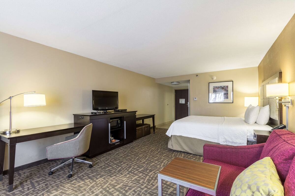 A hotel room with a bed, a desk with a computer, a chair, a television, and a small table.