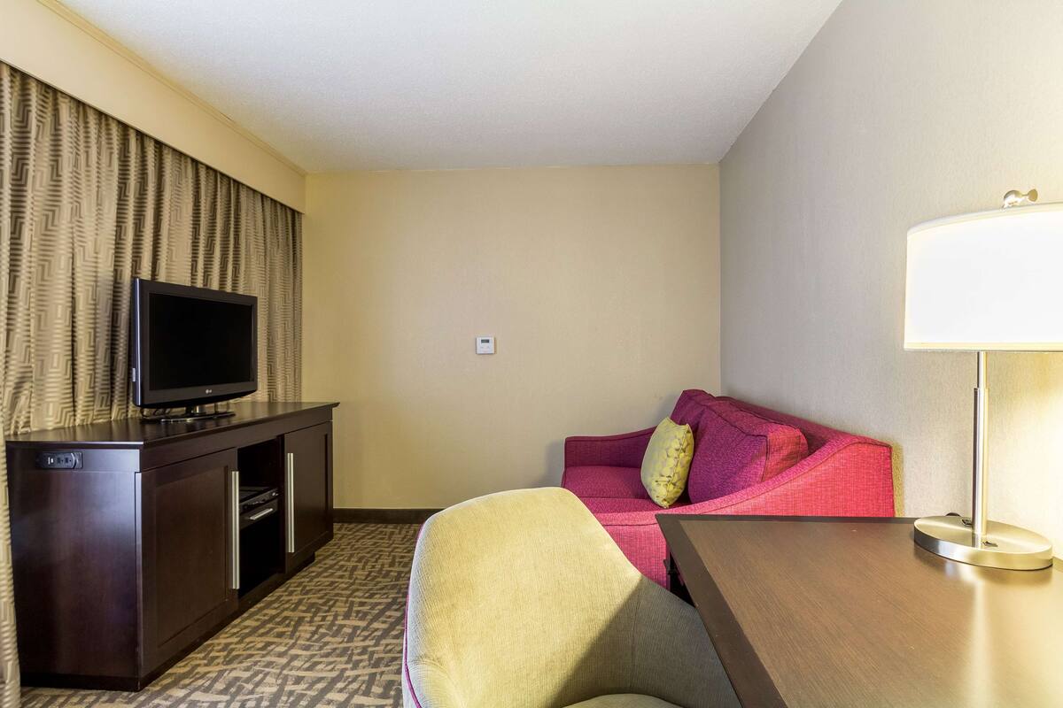 A hotel room with a flat-screen TV on a dark wooden cabinet, a red and yellow sofa, a small table, and a floor lamp.