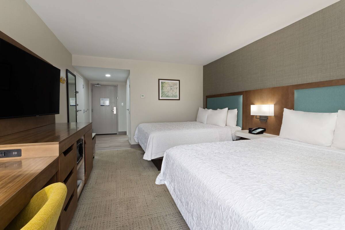 A hotel room with two beds, a desk with a flat-screen TV, a wall-mounted lamp, and a framed picture on the wall.