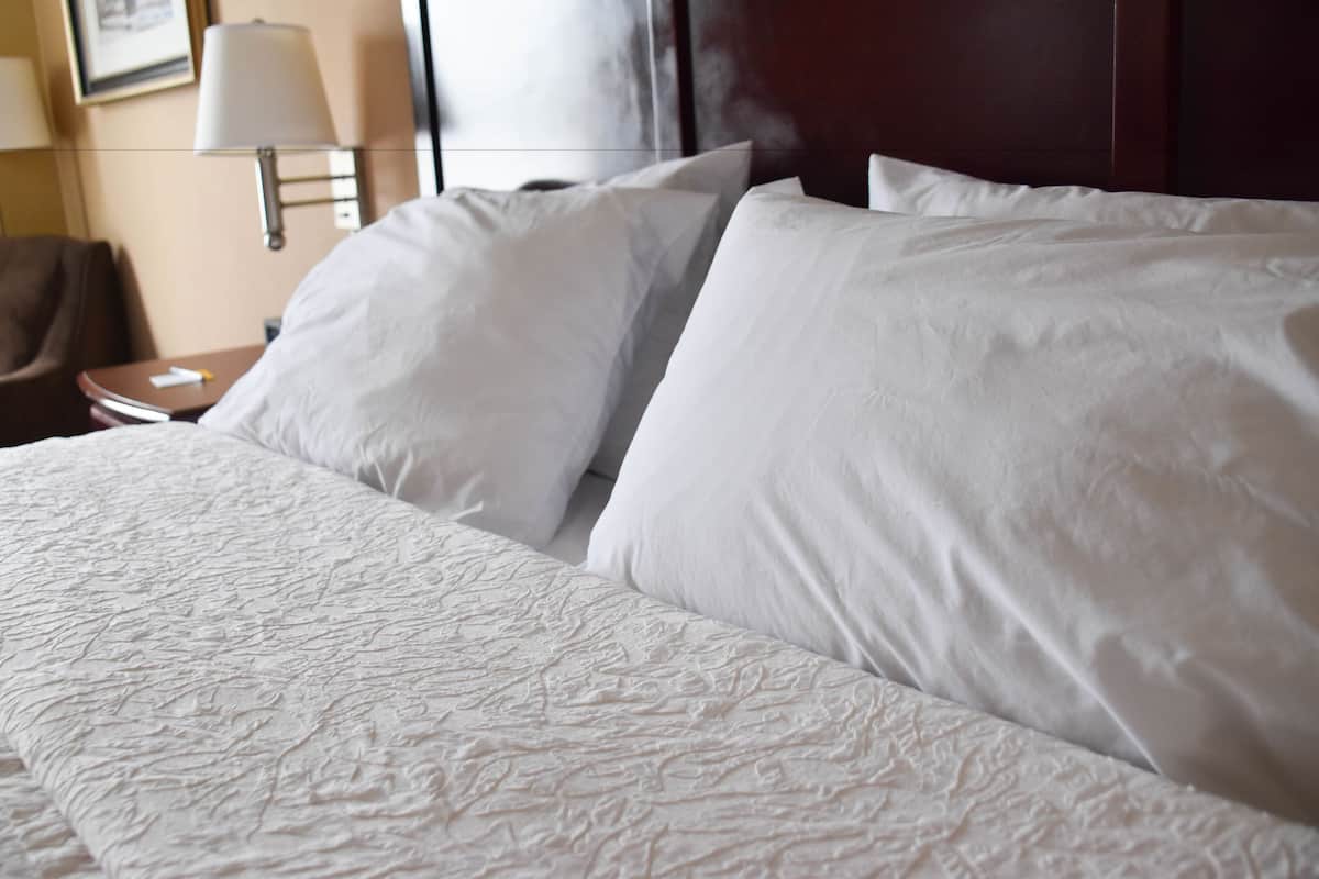 A neatly made bed with white linens and a textured duvet cover, a bedside table with a lamp, and a framed picture on the wall.