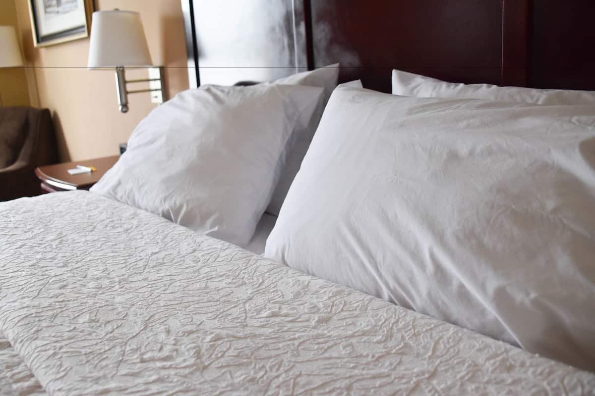 A neatly made bed with white linens, a wooden headboard, and a nightstand with a lamp.