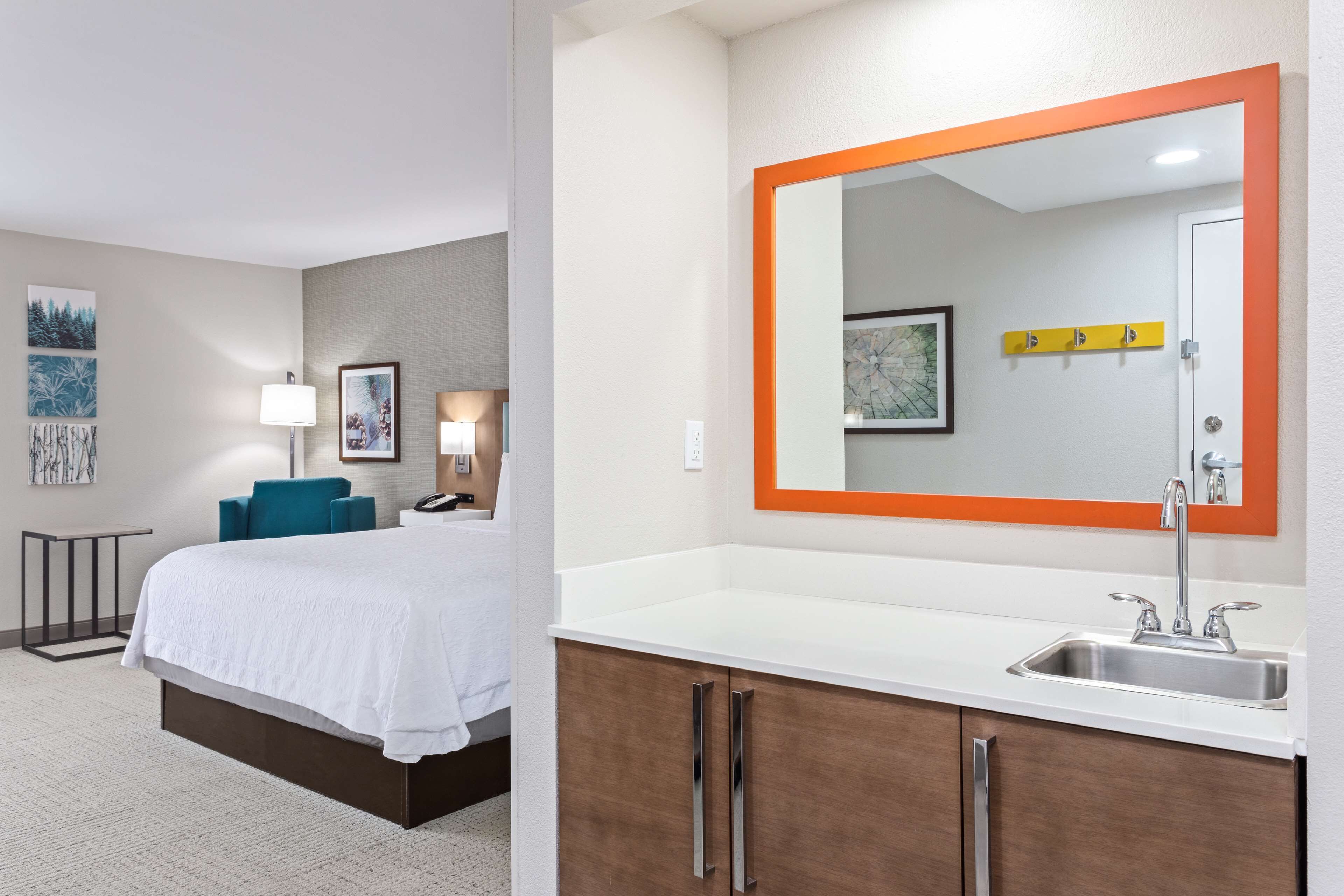Habitación de hotel con cama, mesita de noche, lámpara y un espejo grande sobre un lavabo blanco de diseño moderno.