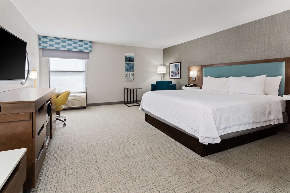 A hotel room with a large bed, a desk with a chair, a TV, and a window with blinds.