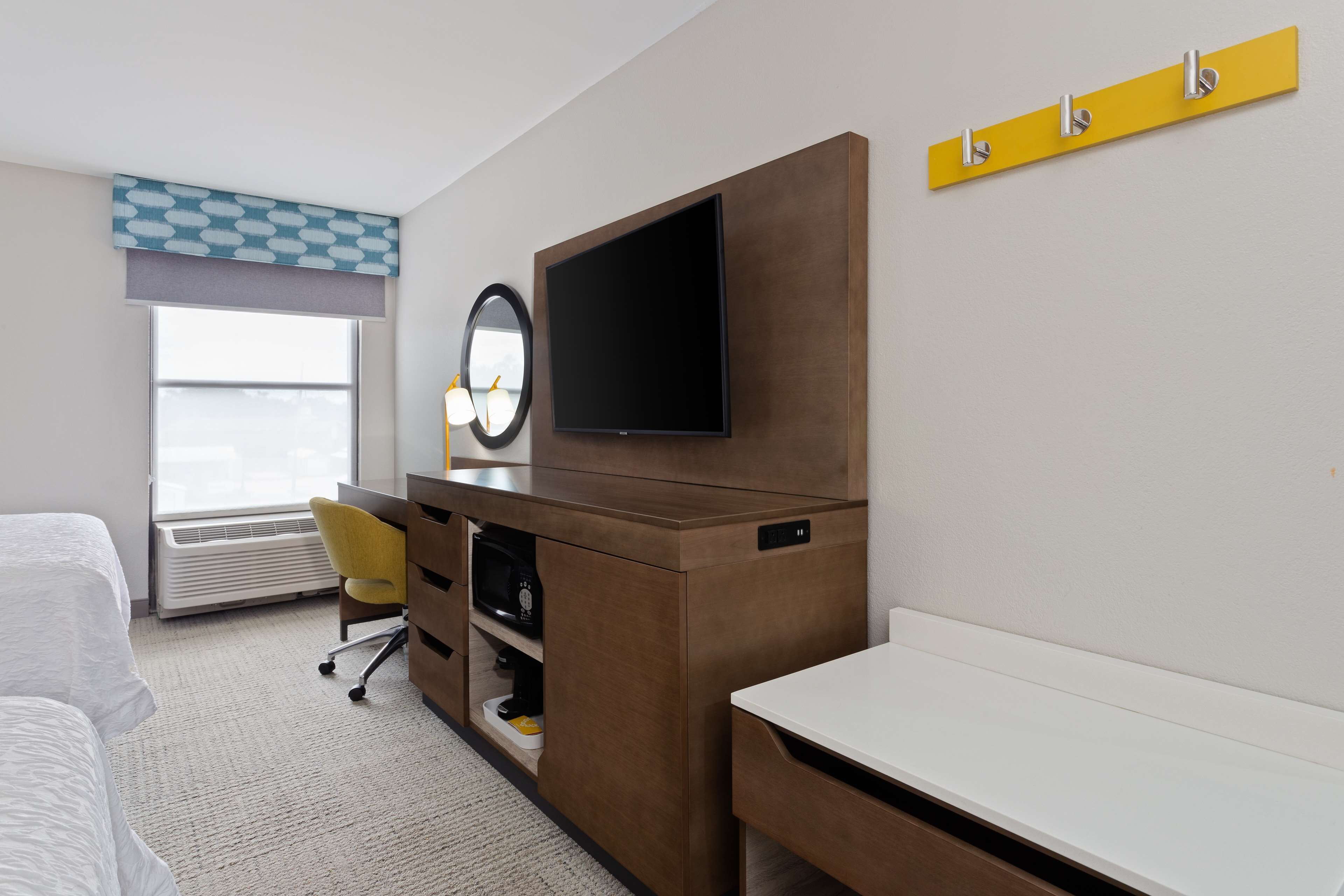 Habitación de hotel con cama, escritorio, silla, televisor, espejo y ventana con persianas.