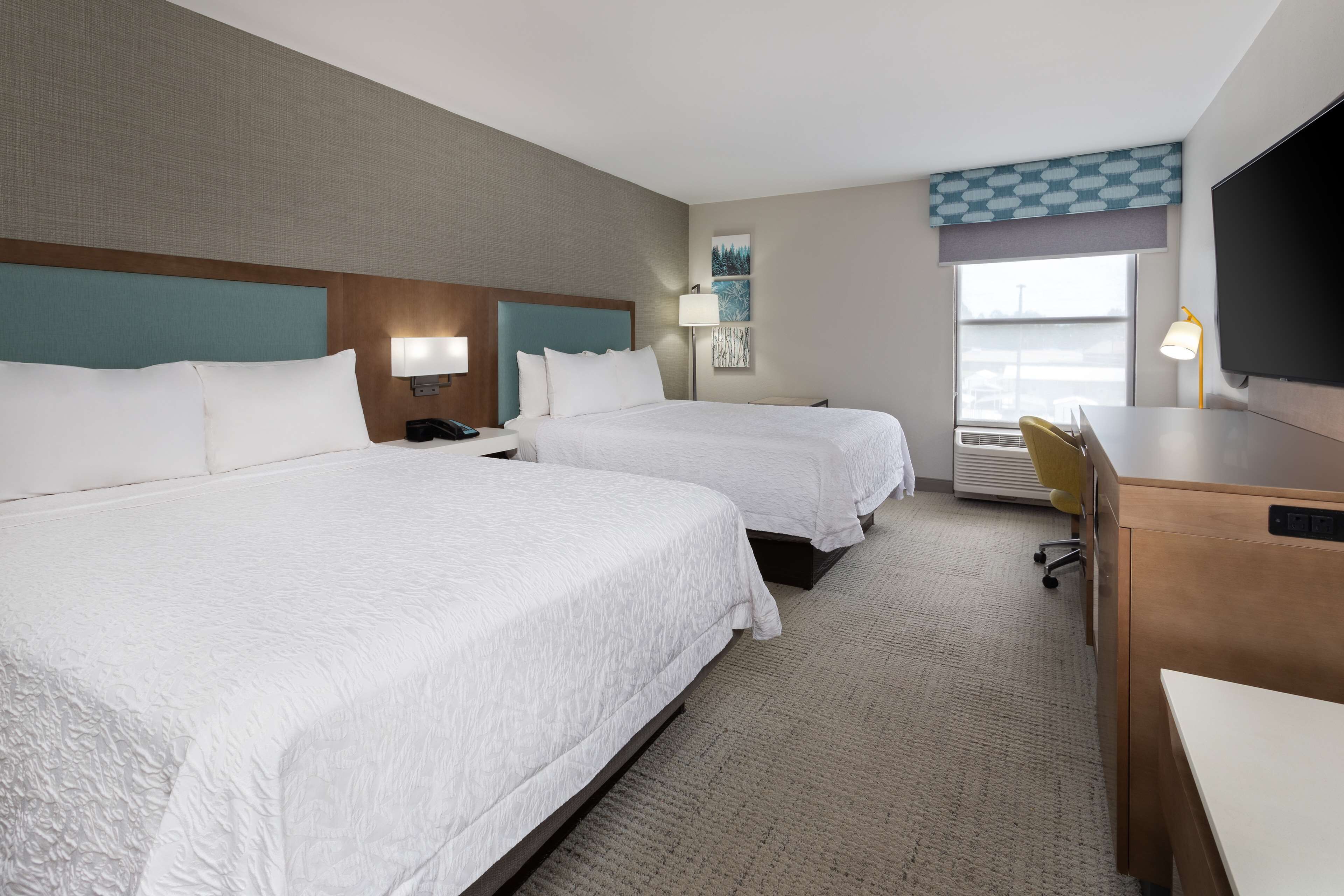 A hotel room with two beds, a desk, a chair, a flat-screen TV, a lamp, and a window with a view.