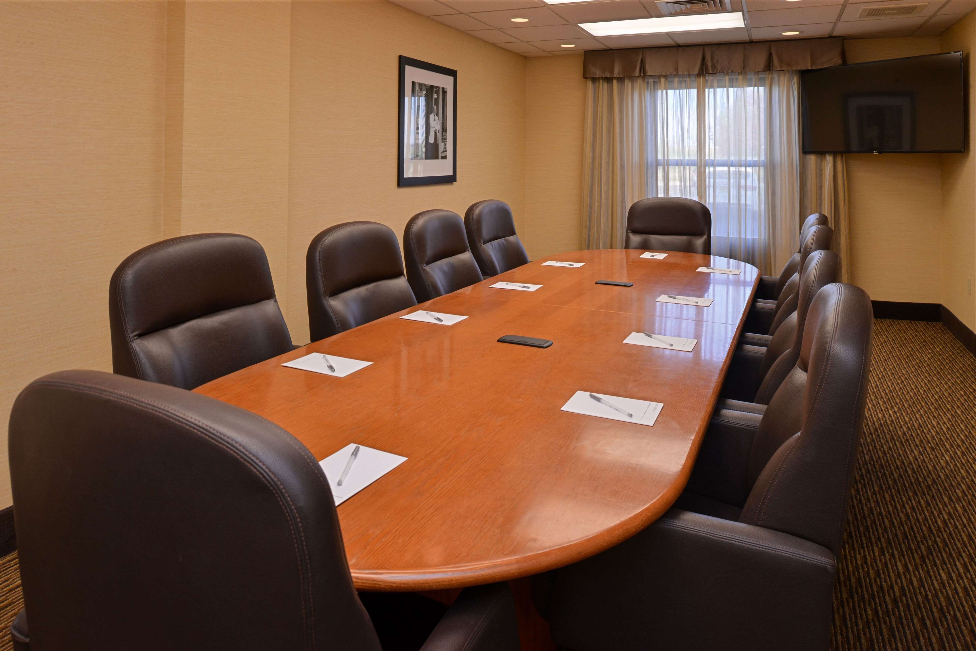 A conference room with a long table and eight chairs, a framed picture on the wall, and a TV mounted above the table.