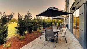 An outdoor patio with a table and chairs, a large umbrella, and a view of a grassy area and trees.