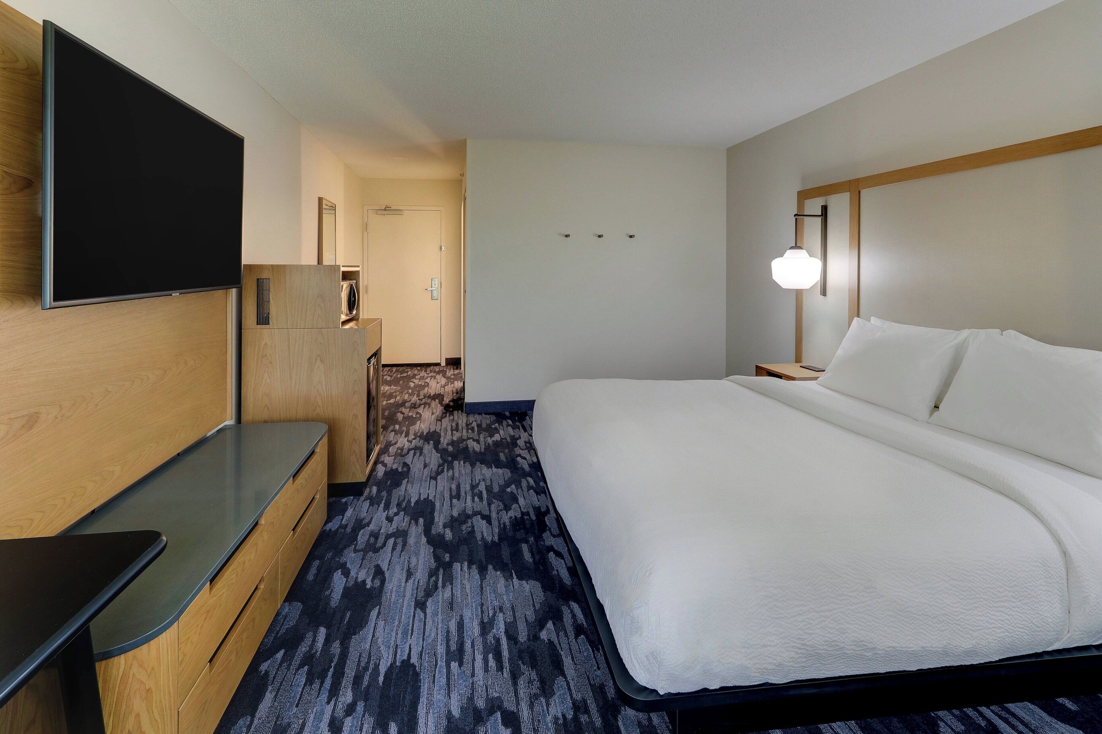 A hotel room with a bed, a desk, a flat-screen TV mounted on the wall, and a carpeted floor.
