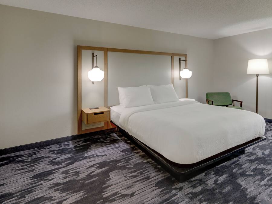 Una habitación de hotel con una cama grande, una mesita de noche, una silla verde y dos lámparas de pared.
