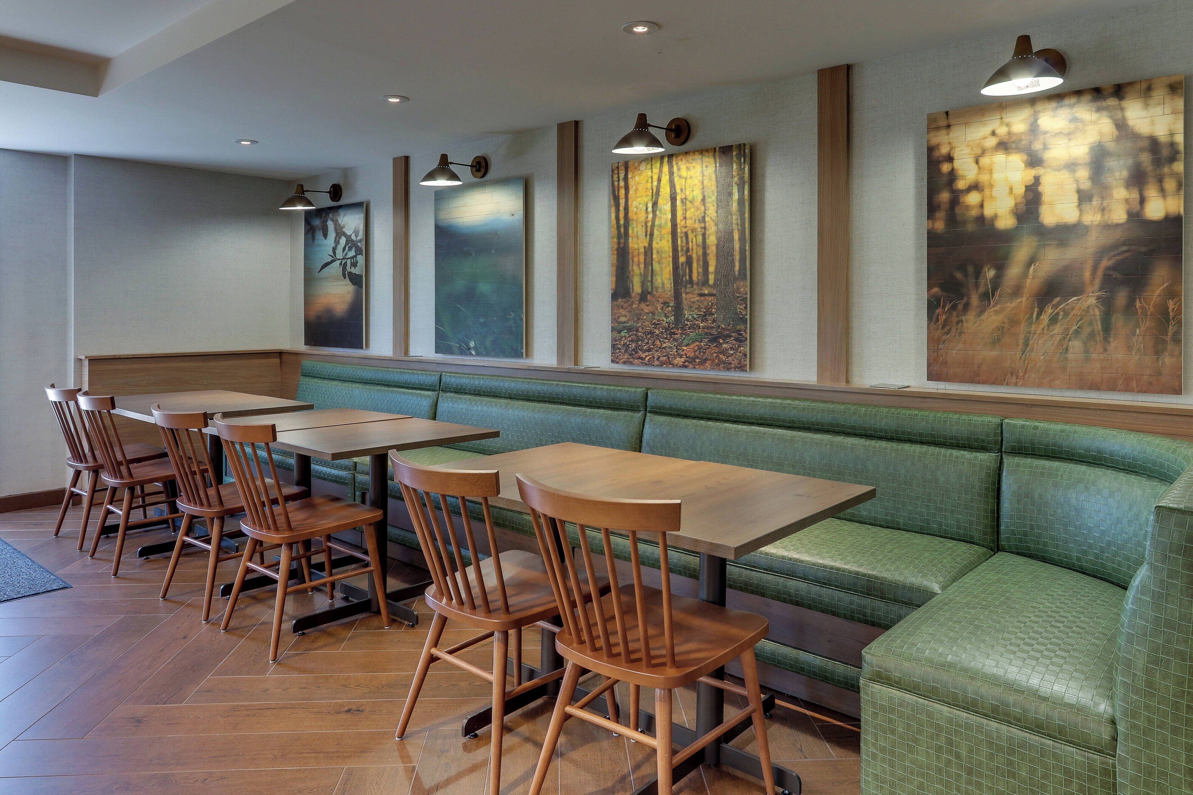 Interior de un restaurante con mesas y sillas de madera, asientos acolchados verdes y obras de arte en las paredes.