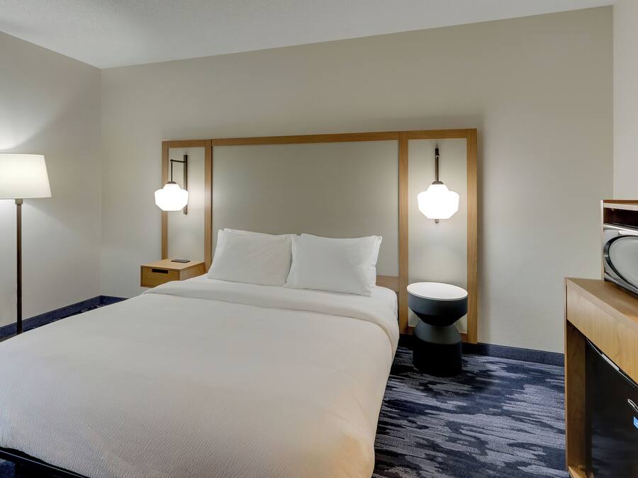 Ett hotellrum med en stor säng, sänglampor, en sänggavel i trä, ett litet nattduksbord, ett runt svart sidobord och en mikrovågsugn placerad på ett träskåp.