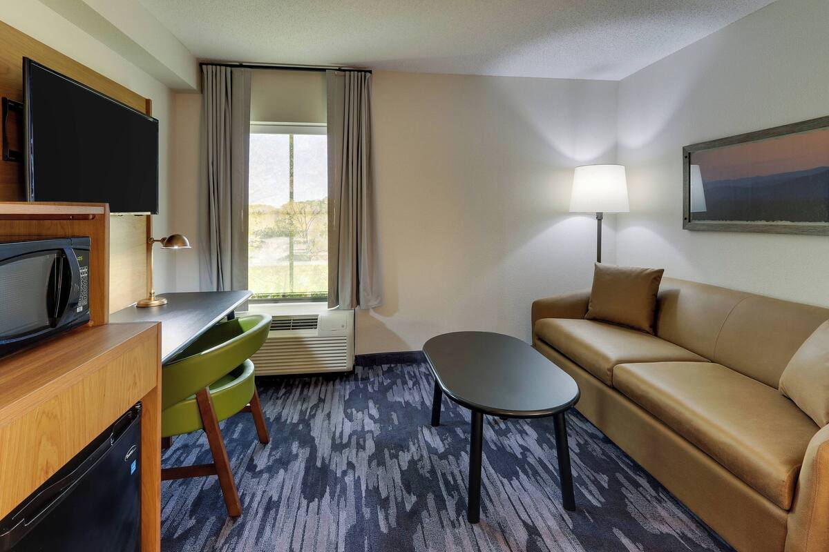 Een hotelkamer met een bank, een kleine tafel, een televisie, een bureau met stoel en een raam met gordijnen.